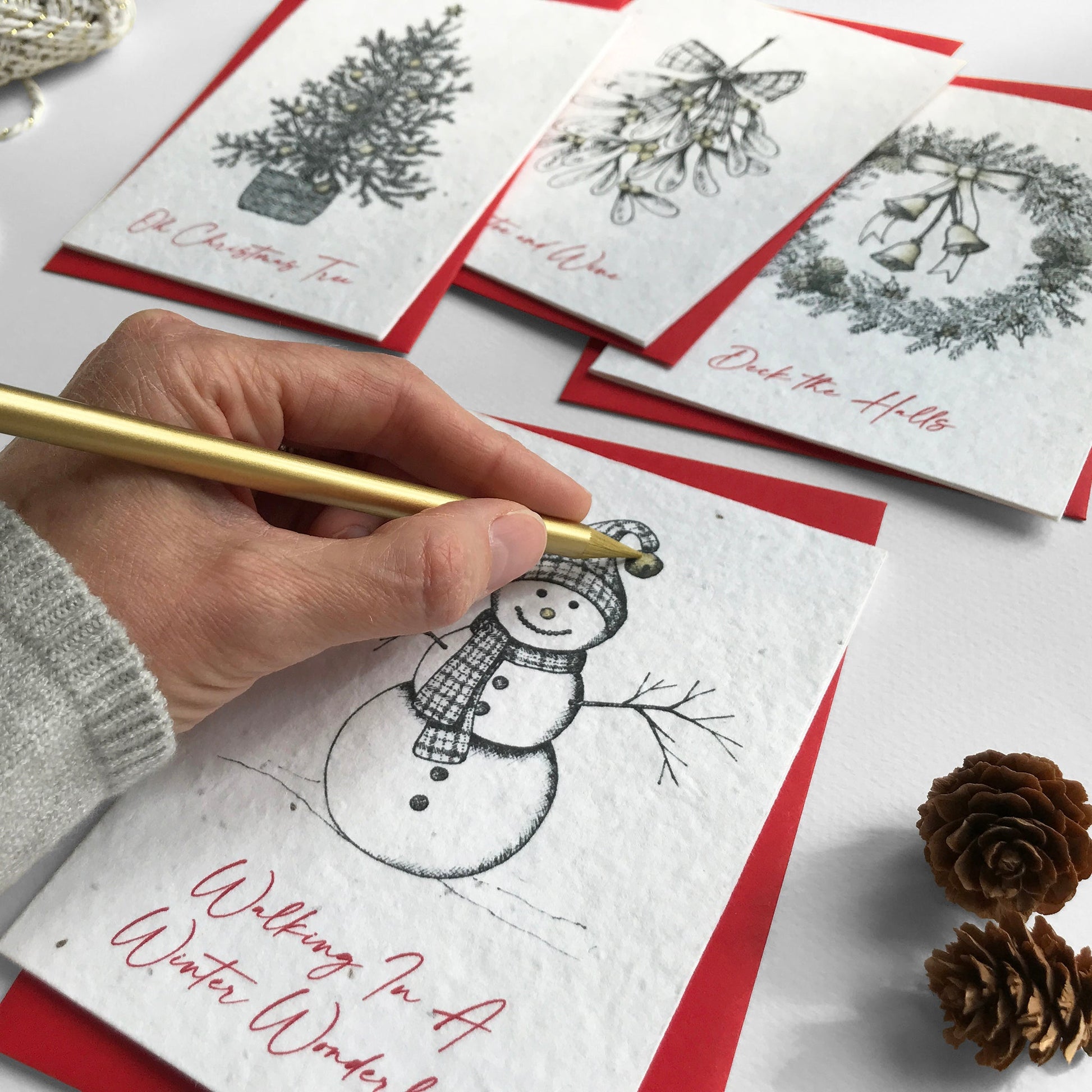 Image shows a pack of A6 plantable Festive Illustration Christmas cards being hand finished with gold pencil. There are 4 card designs in the pack, each with a recycled red envelope. The hand illustrated cards are printed onto plantable seed paper so when the festivities are over, they can be planted to grow bee friendly flowers for the garden.