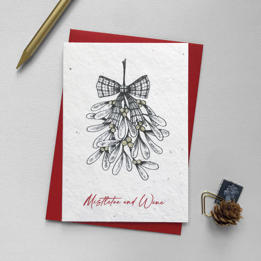 Image shows an A6 plantable Christmas Illustration card featuring pen illustrated Mistletoe. The berries are hand finished using a gold pencil. The red text underneath reads 'Mistletoe and Wine'. This eco-friendly recycled cotton card is embedded with seeds so it can be planted in spring to grow beautiful flowers for the garden. Each biodegradable card comes with a recycled red envelope.