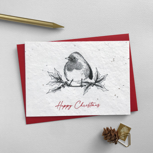 Image shows a Bloom Cards A6 plantable Winter Animals Christmas card featuring a robin sitting on a holly branch. The red script style text underneath reads 'Happy Christmas'. This eco-friendly, biodegradable card is made from recycled cotton embedded with bee friendly wildflower seeds, so when the celebrations are over, it can be planted to grow beautiful flowers for the garden. Each card comes with a recycled red envelope.
