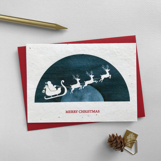 Image shows an A6 Bloom Cards plantable Christmas card featuring a navy blue semi-circular scene with a white silhouette cut-out of Father Christmas and three reindeer flying the sleigh. Rudolph's nose and details on the sleigh have been hand finished using a gold pencil. The red text underneath reads 'Merry Christmas'. This recycled card can be planted to grow a mix of sweet alyssum, poppy, baby's breath and rudbeckia flowers. Each card comes with a recycled red envelope.