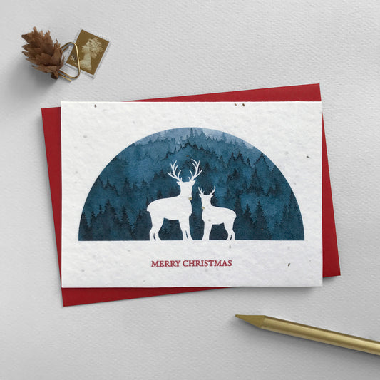 Image shows an A6 Bloom Cards plantable Christmas card featuring a navy blue semi-circular scene with a white silhouette cut-out of two reindeer. The noses of the reindeer are hand finished using a gold pencil. The red text underneath reads 'Merry Christmas'. Plant this eco-friendly card can be planted to grow a mix of sweet alyssum, poppy, baby's breath and rudbeckia flowers. Each card comes with a recycled red envelope.