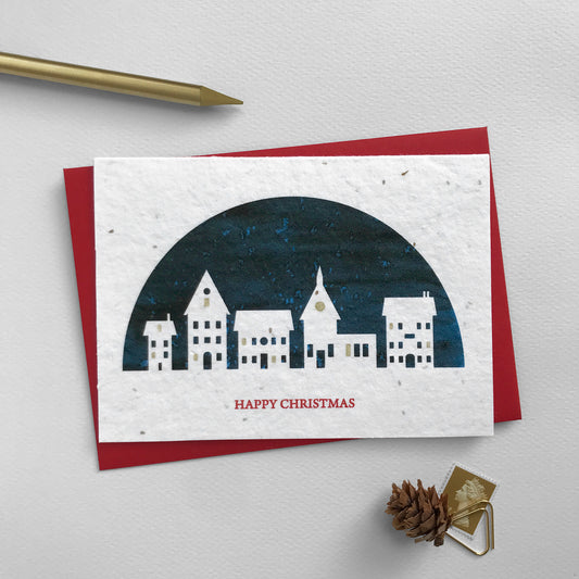 Image shows an A6 Bloom Cards plantable Christmas card featuring a navy blue semi-circular scene with a white silhouette cut-out of houses and a church. Some of the windows are hand finished using a gold pencil giving the appearance of lights being on in the window. The red text underneath reads 'Happy Christmas'. Plant this eco-friendly card to grow a mix of sweet alyssum, poppy, baby's breath and rudbeckia. Each card comes with a recycled red envelope.
