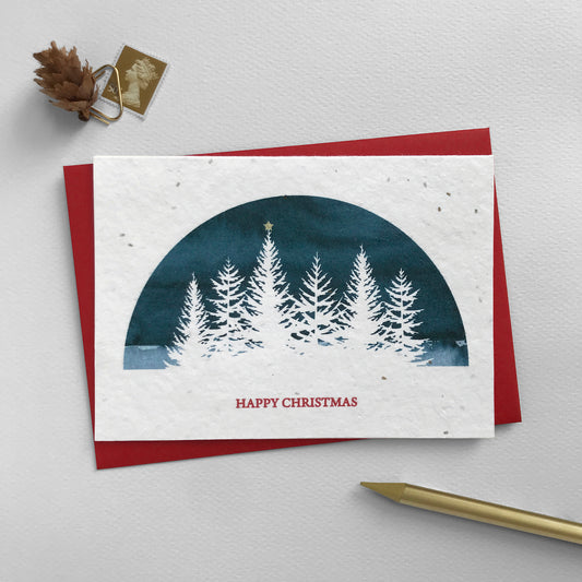 Image shows an A6 Bloom Cards plantable Christmas card featuring a navy blue semi-circular scene with a white silhouette cut-out fir trees, one with a star on top. The star has been hand finished using gold pencil. The red text underneath reads 'Happy Christmas'. Plant this eco-friendly card to grow a mix of sweet alyssum, poppy, baby's breath and rudbeckia flowers. Each card comes with a recycled red envelope.