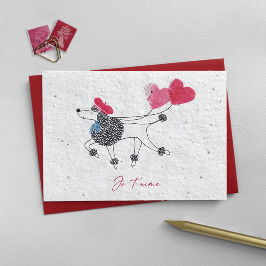 Image shows a Suzanne Marie Paperie plantable wildflower seed valentines card featuring a pen illustrated French Poodle. He is joyfully trotting along wearing a watercolour red beret and blue neckerchief and carrying with him two watercolour heart balloons in pink and red. The red script text underneath reads 'Je t'aime'. This cute card comes with a red recycled envelope and is photographed with red and pink stamps and gold pencil and paperclip.