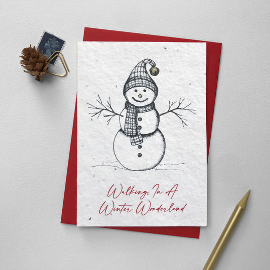 Image shows an A6 plantable Christmas Illustration card featuring pen illustrated snowman with hat and scarf. The snowman's nose and bell on his scarf are hand finished using a gold pencil. The red text underneath reads 'Walking in a Winter Wonderland'. This eco-friendly, biodegradable card is embedded with seeds so it can be planted to grow beautiful flowers for the garden. Each card comes with a recycled red envelope.