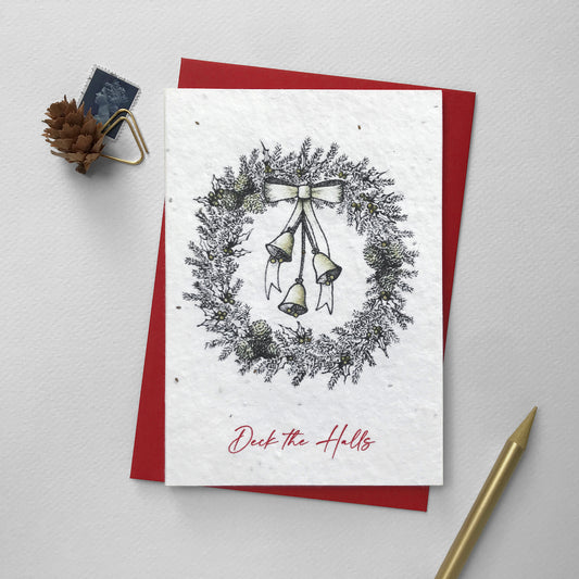 Image shows an A6 plantable Christmas Illustration card featuring pen illustrated Christmas Wreath with bells, bow, berries and pine cones. The bells, bow, berries and pine cones are hand finished using a gold pencil. The red text underneath reads 'Deck the Halls'. This eco-friendly recycled cotton card is embedded with seeds so it can be planted to grow beautiful wildflowers for the garden. Each card comes with a recycled red envelope.