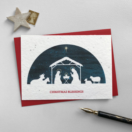 Image shows a Bloom Cards A6 plantable Christmas card featuring a navy blue semi-circular scene with a white silhouette cut-out of the nativity scene with Mary, Joseph, baby Jesus, and cattle. Halos on Mary, Joseph and Jesus along with the star of Bethlehem are hand finished in gold pencil. Red text underneath reads 'Christmas Blessings'. This eco-friendly card can be planted to grow wildflowers for the garden. Each card comes with a recycled red envelope.