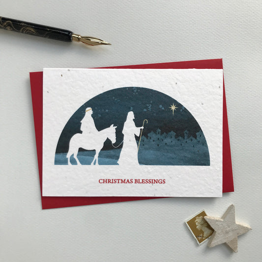 Image shows a Bloom Cards A6 plantable Christmas card featuring a navy blue semi-circular scene with a white silhouette cut-out of Mary, travelling by donkey, with Joseph as they follow the star to Bethlehem. Mary's veil, Joseph's crook and the star of Bethlehem are hand finished in gold pencil. Red text underneath reads 'Christmas Blessings'. Plant this eco-friendly card to grow bee friendly Wildflowers. Each card comes with a recycled red envelope.