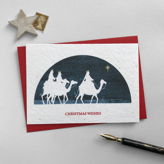 Image shows a Bloom Cards A6 plantable Christmas card featuring a navy blue semi-circular scene with a white silhouette cut-out of the three wise men riding camels and following the star to Bethlehem. Their crowns and the star of Bethlehem are hand finished in gold pencil. Red text underneath reads 'Christmas Wishes'. This eco-friendly card can be planted to grow bee friendly wildflowers. Each card is blank inside and comes with a recycled red envelope.