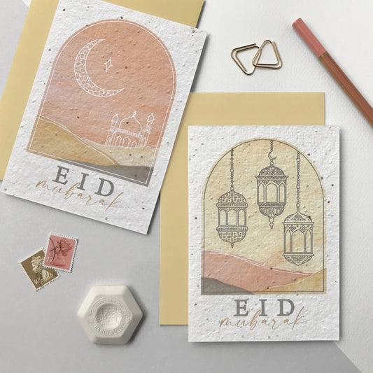 Image shows two wildflower seed Eid greetings cards. The cards feature arch shape designs with watercolour landscapes in pale terracotta and natural. One card features a white line drawing of a moon and mosque and the other with a grey line drawing of three lanterns. The words Eid Mubarak sit underneath. The cards come with matching natural coloured recycled envelopes.
