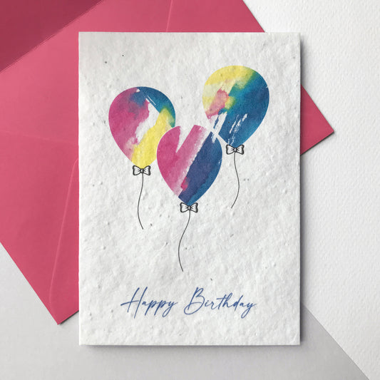 Image shows a Bloom Cards plantable birthday card featuring a hand illustrated watercolour and pen design of three bright multicoloured balloons in blue, pink and yellow. The blue text underneath reads 'Happy Birthday. The card is blank inside and comes with a magenta pink fsc certified eucalyptus envelope. Plant this eco-friendly wildflower seed card and grow a beautiful mix of sweet alyssum, rudbeckia, babies breath and poppies.