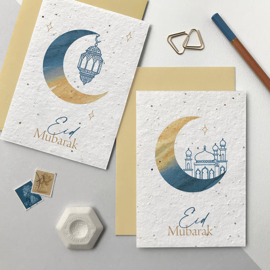Photo of a pack of two plantable Eid greetings cards featuring a watercolour crescent moon in blue and natural. One moon has a blue pen illustrated hanging lantern illustration and the other a blue pen illustrated mosque. The blue and natural text on the cards reads Eid Mubarak. The cards come with natural colour recycled envelopes.