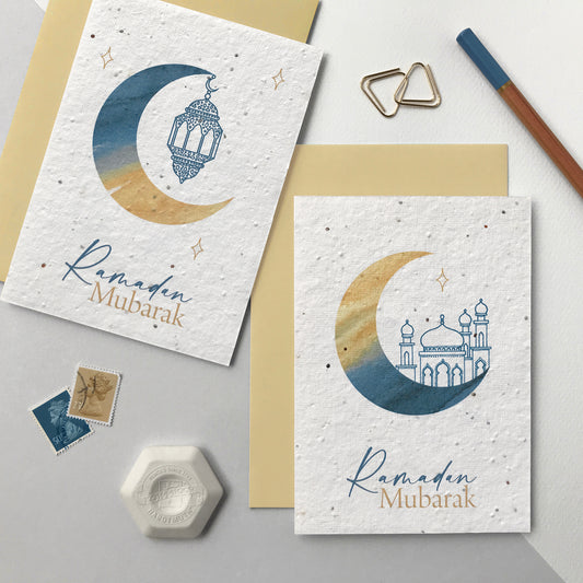 Photo of a pack of two plantable Ramadan greetings cards featuring a watercolour crescent moon in blue and natural. One moon has a blue pen illustrated hanging lantern illustration and the other a blue pen illustrated mosque. The blue and natural text on the cards reads Ramadan Mubarak. The cards come with natural colour recycled envelopes.