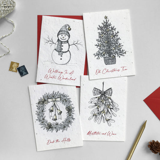 Image shows a pack of plantable seed paper Festive Illustration cards. Each pen illustrated card is hand finished with gold pencil detail and has red script style text. Snowman card reads 'Walking in a winter wonderland'. Christmas Tree card reads 'Oh Christmas Tree'. Wreath card reads 'Deck the Halls'. Mistletoe card reads 'Mistletoe and Wine'. Printed onto recycled paper embedded with seeds, simply plant the card and grow flowers for the garden! Cards are blank inside and come with recycled red envelopes.