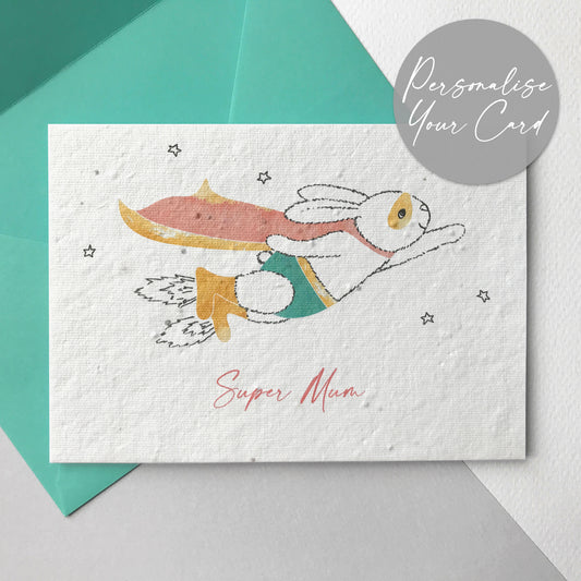 A photo of a hand illustrated Super Mum plantable wildflower seed card in mint. A pen drawn bunny is flying through the stars wearing a watercolour painted coral pink and yellow cape, mint green pants, yellow mask, belt and rocket boots. The coral pink personalised text underneath reads 'Super Mum'. The card comes with a mint green fsc certified eucalyptus paper envelope.