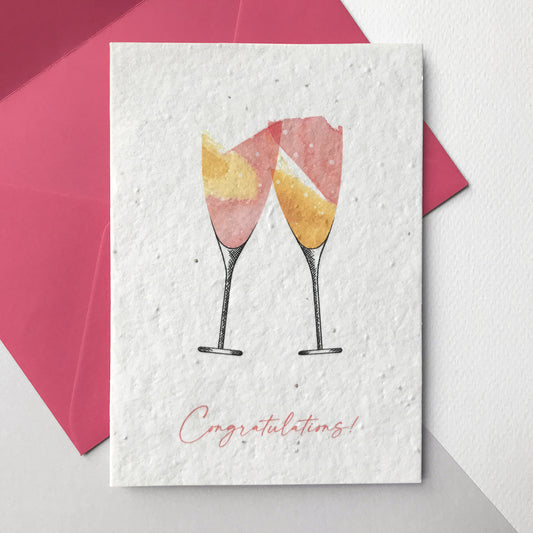Image shows a Bloom Cards A6 plantable seed paper card with a pair of hand illustrated champagne glasses in washes of pink and yellow. The pink text underneath reads 'Congratulations'. These eco-friendly recycled cotton cards are embedded with wildflower seeds so they can be planted to grow beautiful flowers. Each card is blank inside and comes with a magenta fsc certified eucalyptus envelope.
