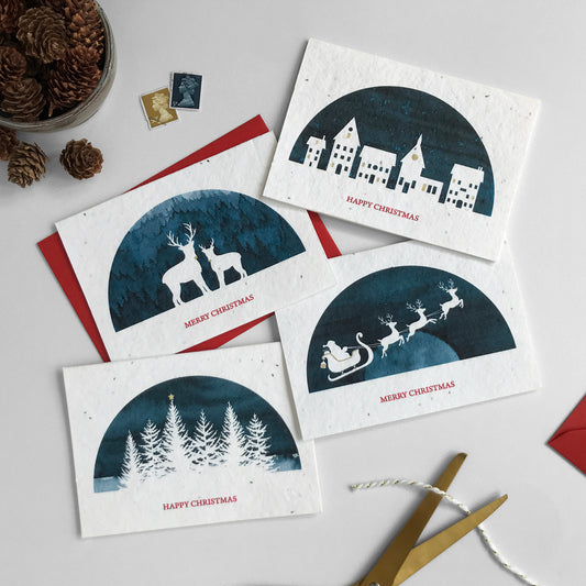 Image shows a pack of Bloom Cards plantable Christmas Scene cards. The cards feature four navy blue watercolour designs with white silhouette cut outs of reindeer, houses, trees and Father Christmas flying his sleigh. Each card is hand finished with gold pencil details. The text underneath reads 'Happy Christmas' and 'Merry Christmas'. Printed onto recycled paper embedded with seeds, plant your card and grow Wildflowers! Cards are blank inside and come with matching recycled red envelopes.