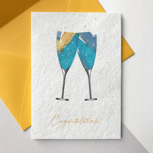 Image shows a Bloom Cards A6 plantable seed paper card with a pair of hand illustrated champagne glasses in washes of turquoise and yellow. The yellow text underneath reads 'Congratulations'. These eco-friendly recycled cotton cards are embedded with wildflower seeds, so they can be planted to grow beautiful flowers for the garden or patio. Each card is blank inside and comes with a yellow fsc certified eucalyptus envelope. Lovingly handmade in Lancashire by Suzanne Marie Paperie.