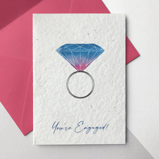 Image shows a Bloom Cards engagement card with a hand illustrated engagement ring design. A bold blue and magenta pink watercolour diamond sits on a pen drawn ring. The blue text underneath reads 'You're Engaged!' Each card is printed onto recycled cotton paper embedded with a choice of bee friendly wildflower, daisy or celosia seeds so the happy couple can enjoy their card then plant it to grow beautiful flowers. The card is blank inside and comes with a magenta pink fsc certified eucalyptus envelope.