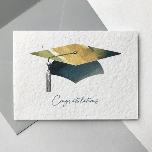 Image shows a Bloom Cards A6 plantable graduation card featuring a watercolour mortar board design with colourful washes of grey, green and natural and a pen drawn tassel. The grey text underneath reads 'Congratulations'. The card is printed onto recycled cotton paper embedded with wildflower seeds so it can be planted to grow beautiful bee friendly flowers. The card is blank inside and comes with a grey fsc certified eucalyptus paper envelope.