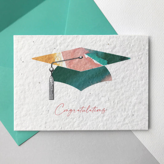 Image shows a Bloom Cards A6 graduation card printed onto plantable seed paper. This eco-friendly card is made from recycled waste cotton and can be planted to grow bee friendly wildflowers. The hand illustrated graduation hat design features colourful washes of coral, mint and yellow and a pen drawn tassel. The pink text underneath reads 'Congratulations'. The card is blank inside and comes with a mint green fsc certified eucalyptus paper envelope. Made by Suzanne Marie Paperie.