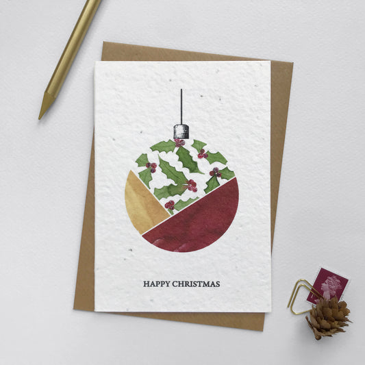 Image shows an A6 Bloom Cards plantable Christmas card featuring a hand illustrated  bauble with a holly design. The holly berries have been hand finished with Bio-glitter Pure. This plastic free glitter is proven to biodegrade in the natural environment in around 4 weeks. The black text underneath reads 'Happy Christmas'. Printed onto recycled paper embedded with Wildflower seeds, simply plant the card to grow beautiful flowers for the garden. Comes with a recycled kraft envelope.