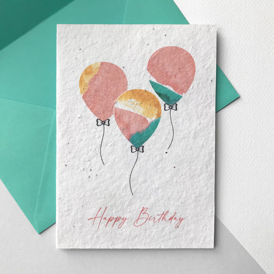 Image shows a Bloom Cards plantable birthday card featuring a hand illustrated watercolour and pen design of three bright multicoloured balloons in mint, coral and yellow. The coral text underneath reads 'Happy Birthday. The card is blank inside and comes with a mint green fsc certified eucalyptus envelope. Plant this seed paper card and grow beautiful bee friendly wildflowers. Made in Lancashire by Suzanne Marie Paperie..