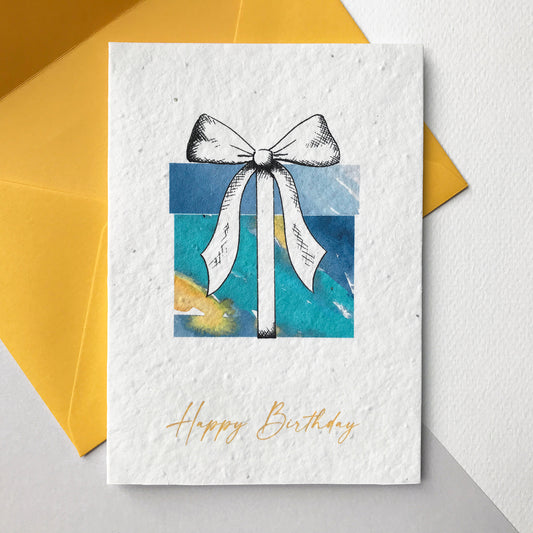 Image shows a Bloom Cards A6 plantable card featuring a brightly coloured blue, turquoise and yellow watercolour present with a pen drawn bow. The yellow text underneath reads 'Happy Birthday'. The hand illustrated design is printed onto eco-friendly recycled cotton paper embedded with seeds so that it can be planted to grow bee friendly wildflowers. The card is blank inside and comes with a matching yellow fsc certified eucalyptus paper envelope.