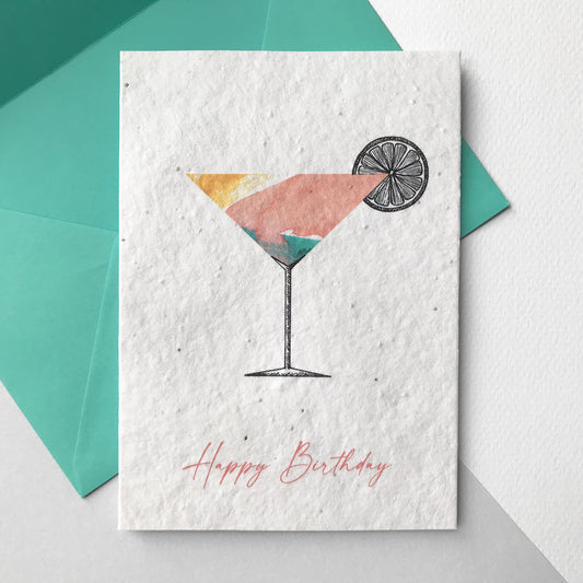 Image shows a Bloom Cards A6 plantable birthday card with a hand illustrated cocktail design. Watercolour washes in yellow, pink and mint make up a colourful cocktail with a pen illustrated stem and slice of lemon. The pink text underneath reads 'Happy Birthday'. Each recycled cotton card is embedded with wildflower seeds so the card can be planted to grow bee friendly flowers for the garden or pots. The card is blank inside and comes with a mint fsc certified eucalyptus envelope.