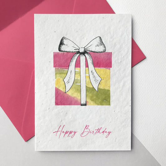 Image shows a Bloom Cards A6 plantable card by Suzanne Marie Paperie featuring a brightly coloured pink, yellow and green watercolour present with a pen drawn bow. The pink text underneath reads 'Happy Birthday'. The hand illustrated design is printed onto eco-friendly recycled cotton paper embedded with seeds so that it can be planted to grow bee friendly wildflowers. The card is blank inside and comes with a matching magenta pink fsc certified eucalyptus paper envelope.
