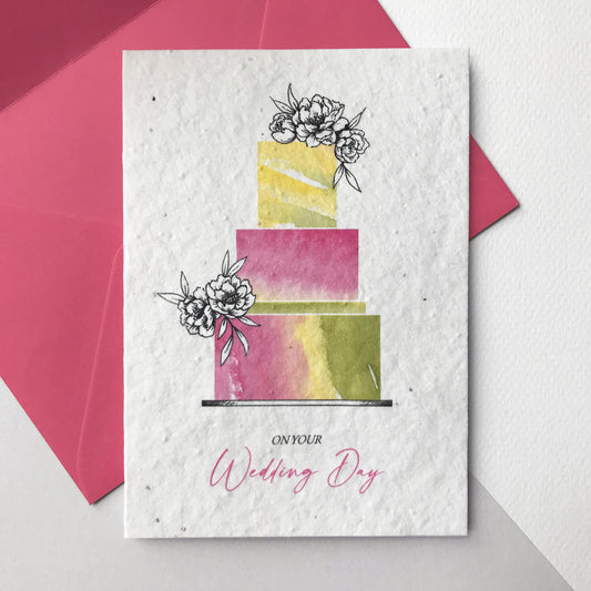 Image of a Bloom Cards A6 plantable wedding card featuring a hand illustrated wedding cake design. Watercolour washes in magenta, green and yellow create a colourful cake with pen drawn flower decoration. The pink text underneath reads 'On Your Wedding Day'. Printed onto eco-friendly paper made from recycled cotton and embedded with wildflower seeds. The card comes with a matching magenta fsc certified eucalyptus envelope. Illustrated and printed in Lancashire by Suzanne Marie Paperie.