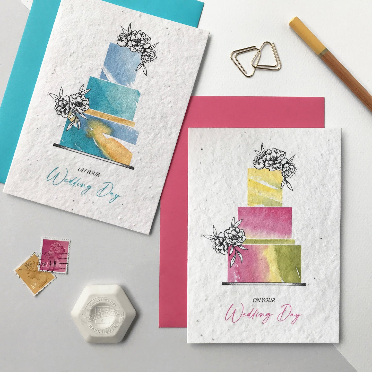 Image shows two Bloom Cards A6 plantable wedding cards in blue and magenta. The cards feature a hand illustrated watercolour and pen wedding cake design. The text underneath reads 'On Your Wedding Day'. Printed onto recycled cotton paper and embedded with wildflower seeds. Each card is blank inside and comes with matching fsc certified eucalyptus paper envelope. Illustrated and printed in Lancashire by Suzanne Marie Paperie.