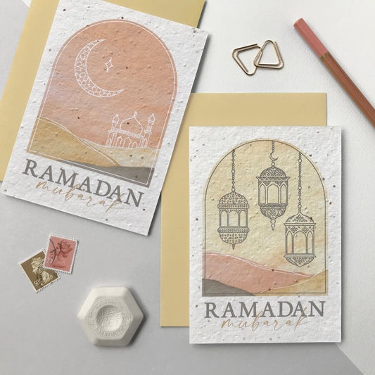 Image shows two wildflower seed Ramadan greetings cards. The cards feature arch shape designs with watercolour landscapes in pale terracotta and natural. One card features a white line drawing of a moon and mosque and the other with a grey line drawing of three lanterns. The words Ramadan Mubarak sit underneath. The cards come with matching natural coloured recycled envelopes.