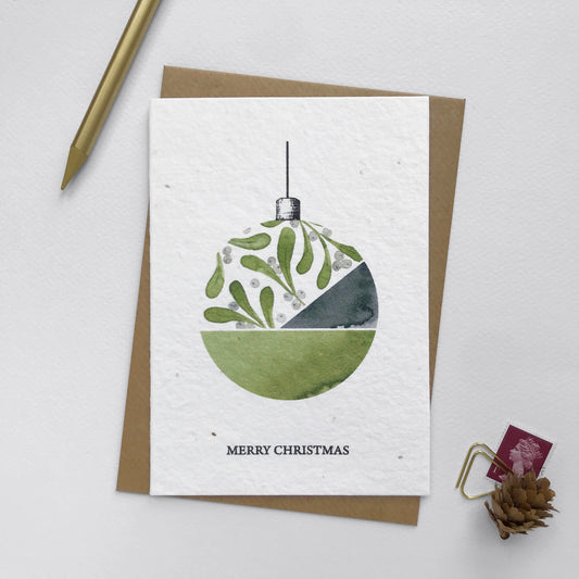 Image shows an A6 Bloom Cards plantable Christmas card featuring a hand illustrated  bauble with a mistletoe design. The berries have been hand finished with Bio-glitter Pure. This plastic free glitter is proven to biodegrade in the natural environment in around 4 weeks. The black text underneath reads 'Merry Christmas'. Printed onto recycled paper embedded with Wildflower seeds, simply plant the card to grow beautiful Wildflowers for the garden. Comes with a recycled kraft envelope.