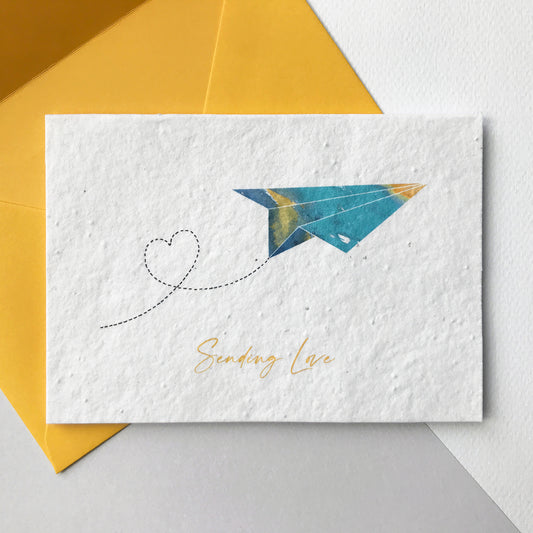 Image of a Bloom Cards A6 plantable note card featuring a hand illustrated design. Watercolour washes in turquoise, blue and yellow are used to create this modern paper plane design with a pen drawn heart trail. The yellow text underneath reads 'Sending Love'. This recycled cotton card is printed onto mwildflower seed paper so your recipient can grow their card and enjoy some beautiful flowers. The card comes with a matching yellow fsc certified eucalyptus envelope. Handmade by Suzanne Marie Paperie.