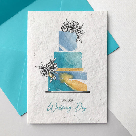 Image of a Bloom Cards A6 plantable wedding card featuring a hand illustrated wedding cake design. Watercolour washes in turquoise, blue and orange create a colourful cake with pen drawn flower decoration. The blue text underneath reads 'On Your Wedding Day'. Printed onto eco-friendly paper made from recycled cotton and embedded with wildflower seeds. The card comes with a matching blue fsc certified eucalyptus envelope.