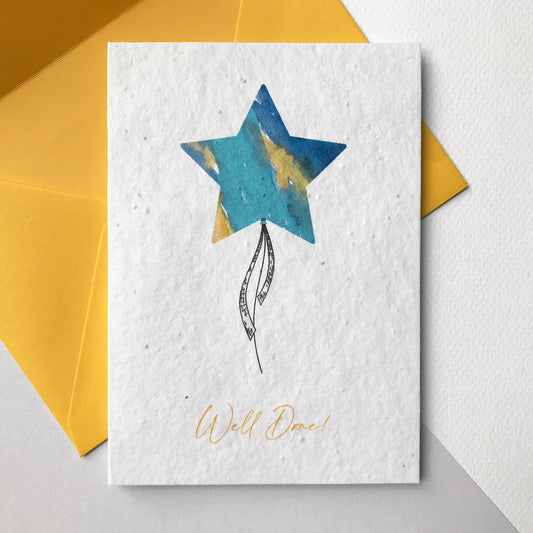 Image shows a Bloom Cards A6 plantable card featuring a brightly coloured turquoise, blue and yellow watercolour star shape balloon with a pen drawn ribbon. The yellow text underneath reads 'Well Done!'. The hand illustrated design is printed onto eco-friendly recycled cotton paper embedded with seeds so that it can be planted to grow bee friendly wildflowers. The card is blank inside and comes with a matching yellow fsc certified eucalyptus paper envelope. Illustrade and printed by Suzanne Marie Paperie.
