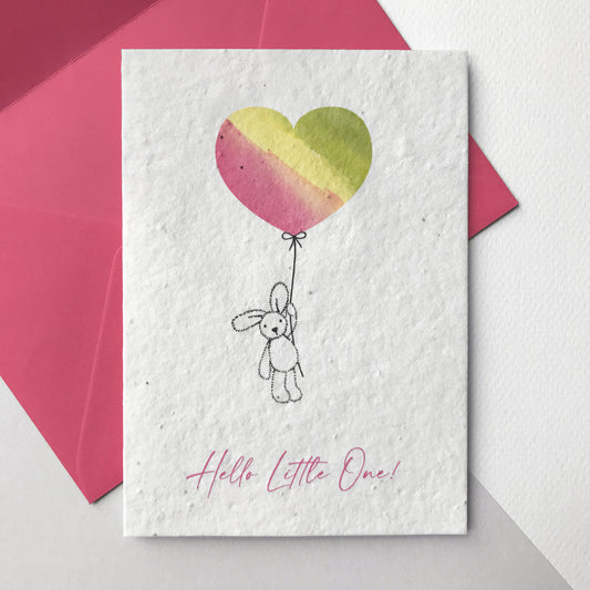 Image of a Bloom Cards A6 plantable New Baby card featuring a cute hand illustrated bunny and balloon design. A pen drawn bunny holds a magenta, yellow and green watercolour heart shape balloon. The magenta pink text underneath reads 'Hello Little One!'. Printed onto eco-friendly paper made from recycled cotton and embedded with wildflower seeds. The card comes with a matching magenta fsc certified eucalyptus envelope. Made in Lancashire by Suzanne Marie Paperie.