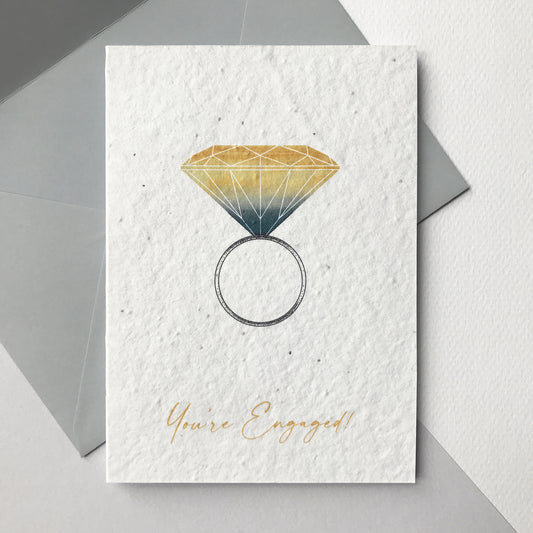 Image shows a Bloom Cards A6 engagement card with a modern hand illustrated engagement ring design. A bold yellow and grey watercolour diamond sits on a pen drawn ring. The yellow text underneath reads 'You're Engaged!' Each card is printed onto recycled cotton paper embedded with bee friendly wildflower seeds so the happy couple can enjoy their card then plant it to grow beautiful flowers. The card is blank inside and comes with a grey fsc certified eucalyptus envelope.