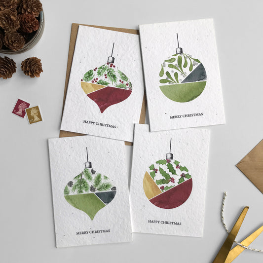 Image shows a pack of A6 Bloom Cards plantable Bauble Christmas cards. The cards feature four hand painted Christmas decorations with fir, mistletoe, pine and holly in festive greens, reds and gold. Each card is hand finished with plastic free, biodegradable glitter. The text underneath reads 'Happy Christmas' and 'Merry Christmas'. Printed onto recycled cotton paper embedded with Wildflower seeds. Cards are blank inside and come with matching recycled kraft envelopes.
